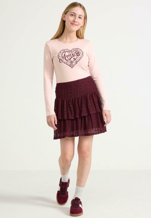Camicia a maniche lunghe rosa chiaro con la scritta "Amore" in un design a cuore. Gonna a balze in pizzo borgogna. Modella indossa sneakers borgogna con calzini grigi.