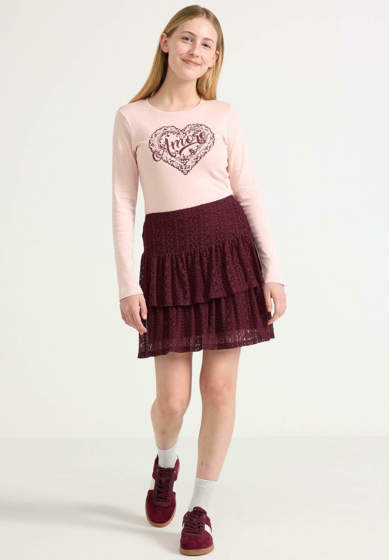 Lichtroze langekle shirts met "Amore" in een hartontwerp. Bordeaurood kanten gelaagd rokje. Model draagt bordeaurode sneakers met grijze sokken.