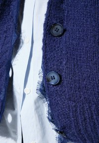 Marine gebreide cardigan met een gestructureerd oppervlak, rafelige randen en twee zwarte knopen, gelaagd over een lichtblauw gestreept shirt.