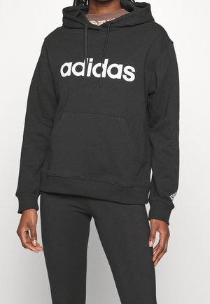 Person iført en sort Adidas hættetrøje med hvidt logo og matchende sorte bukser, stående mod en neutral baggrund.