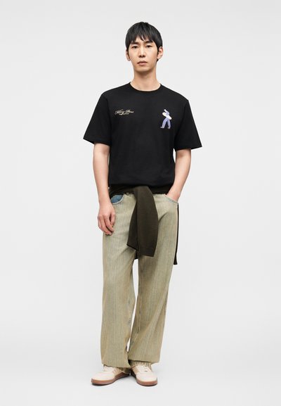 Μαύρο βαμβακερό t-shirt με μικρό γραφικό και χρυσό κείμενο, σε συνδυασμό με ανοιχτό πράσινο παντελόνι με ρίγες και φαρδιά πόδια και καφετιά παπούτσια.