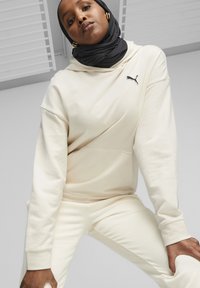 Cremefarvet hoodie med en kængurulomme, der har et lille sort Puma-logo. Sammen med matchende cremefarvede bukser, bæres med en sort hijab.