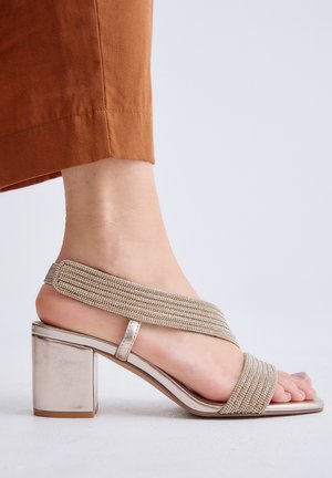 Pie con una sandalia metálica de tacón bloque con dos tiras brillantes, combinado con pantalones recortados de color óxido sobre un fondo claro y liso.