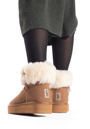 Brown suede ankle boots with white faux fur cuffs and tan rubber soles, featuring logo tags on both sides. Legs in black tights visible.