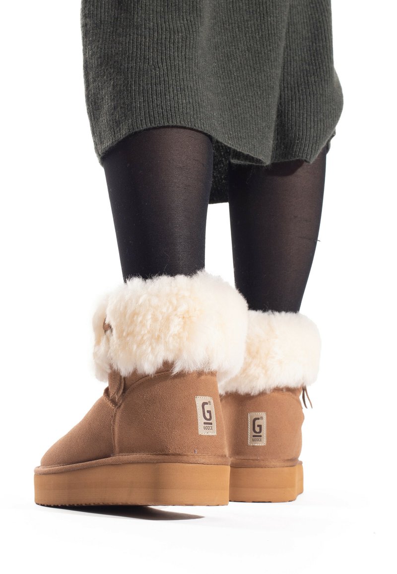 Botas de ante marrón hasta el tobillo con puños de piel sintética blanca y suelas de goma color arena, con etiquetas de logo en ambos lados. Piernas visibles con medias negras.