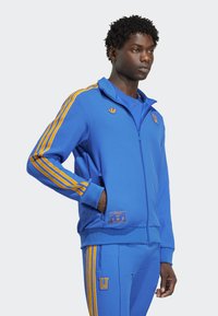 Mand iført en blå zip-up tracksuit med orange striber på ærmerne og bukserne, stående med den ene hånd i lommen og kigger fremad.