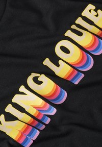 Camiseta de tela negra con el texto "KING LOUIE" en grandes letras redondeadas de colores múltiples con efecto de degradado y colores superpuestos.