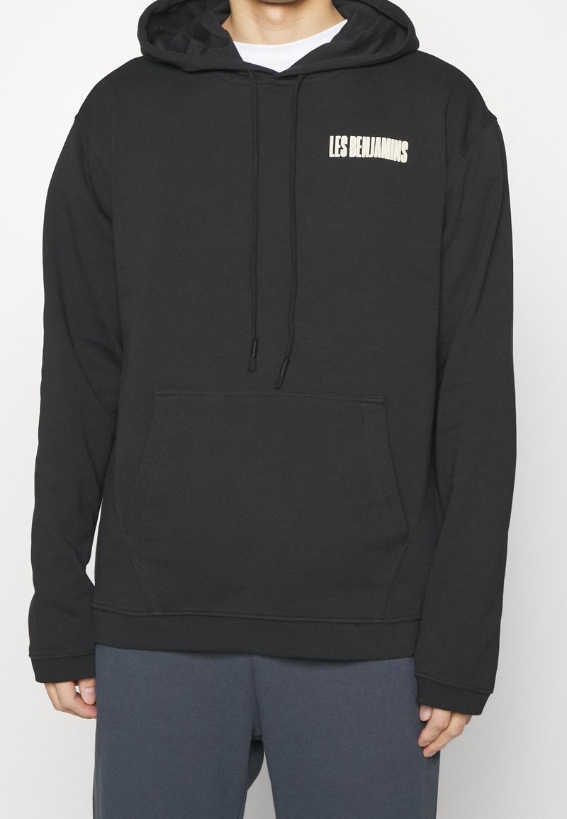 Personne portant un sweat à capuche noir avec le texte « LES BENJAMINS » sur la poitrine et une poche kangourou à l'avant, associé à un pantalon gris foncé.