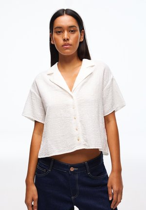 Ženska, oblečena v belo kratkoročno srajco z gumbi s teksturo in temno modre kavbojke, stoji pred enostavnim belim ozadjem.