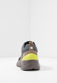 Baskets gris et marron avec une tige en daim et textile, dotées d'un accent jaune au talon et d'une languette marron. Détail subtil du logo à l'arrière.