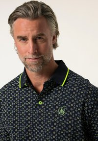 Polo navy con un patrón floral en blanco y verde, con un cuello negro en contraste y acentos verdes en la tapeta y el logo.