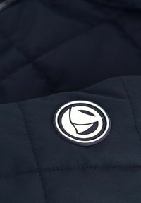 Giacca imbottita blu navy con un logo rotondo in bianco e nero. Il materiale è liscio con una texture trapuntata.