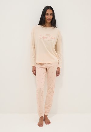 Femme portant un haut beige à manches longues avec le texte "The Coffee Club" et un pantalon de pyjama rose clair à motifs, debout pieds nus devant un fond blanc uni.