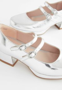 Silberne Mary Jane-Schuhe mit glänzender Metallic-Oberfläche, zwei verstellbaren Riemen, runder Schuhspitze und einem niedrigen Blockabsatz. Glatte Textur und feine Nähte.