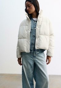 Pufferjakke i creme med en quilted design, parret med lyseblå denimoveralls med knapfront og brede ben.