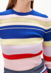 Kvinna som bär en ribbad stickad tröja med horisontella ränder i blått, gult, rosa, rött och grått, visad från axlarna till höfterna.