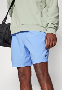 Homme portant un short Nike bleu clair et un sweat-shirt vert clair, tenant un sac de sport noir sur son épaule.