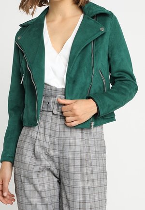 Frau trägt eine grüne Wildleder-Bikerjacke über einem weißen V-Ausschnitt-Oberteil, kombiniert mit hoch taillierten grauen karierten Hosen mit Gürtel.