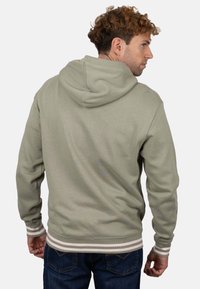 Sudadera con capucha en verde salvia, con puños y dobladillo acanalados que presentan rayas blancas. La tela parece suave y cómoda.