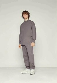Giovane con capelli ricci in piedi, indossa un ampio top grigio a maniche lunghe e pantaloni coordinati con scarpe da ginnastica bianche, su uno sfondo chiaro e semplice.
