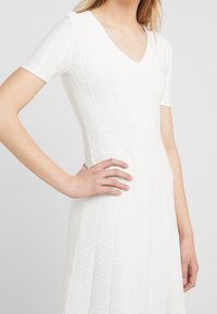 Robe blanche à manches courtes avec un col en V, présentant des rayures texturées et un corsage ajusté qui s'évase en une jupe longueur genou.