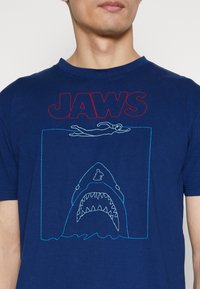 Navyblauwe T-shirt met een grafisch ontwerp met "JAWS" in het rood, een zwemmende figuur omrand in het wit en een haai met gedetailleerde tanden in lichtblauw.