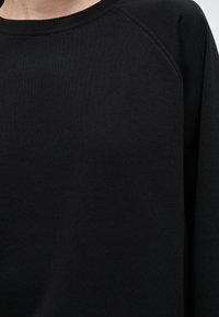 Gros plan sur une personne portant un pull noir uni à col rond avec des détails de coutures visibles à l’épaule et à l’encolure.