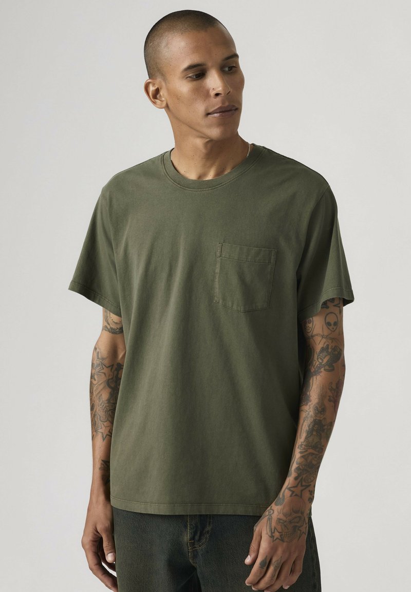 Man met tatoeages op beide armen, gekleed in een olijfgroene T-shirt met zakje en donkere broek, kijkt naar links tegen een effen achtergrond.