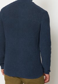 Mörkblå stickad tröja med hög krage, texturerat mönster, ribbade ärmslut och rak fåll. Synliga sömmar på axlarna.