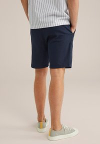 Muž v námořnických modrých šortkách, pruhované tričko vytažené ven a šedé tenisky se žlutým detailem na patě, stojí s rukama v kapsách.