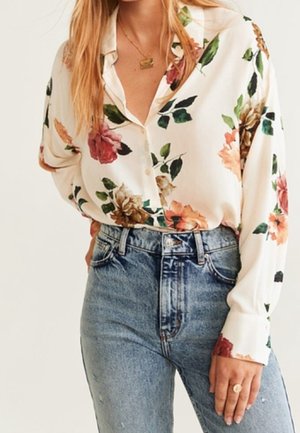 Femme portant un chemisier blanc à manches longues imprimé floral, rentré dans un jean bleu taille haute, avec un collier et une bague en or visibles.