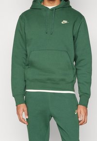 En person som bär en grön Nike-hoodie och matchande gröna sweatpants med vita Nike-loggor, står mot en enkel bakgrund.