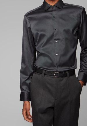 Man wearing a fitted black satin dress shirt tucked into dark gray dress pants with a black belt against a plain background.