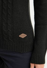 Svart stickad tröja med kabelstickat mönster, ribbade ärmmuddar och en läderlogotyp på nedersta vänstra delen.