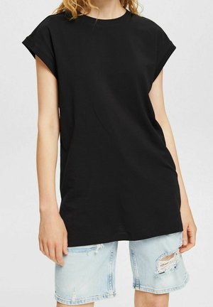 Kvinde iført løs sort ærmeløs t-shirt og lyseblå slidte denimshorts mod en ensfarvet baggrund.