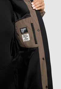 Čierna bunda s hnedým vnútrom, vybavená vreckom na zips, logo nášivkami a hladkou textúrou. Dizajn je funkčný a moderný.