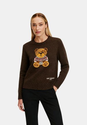 Donna che indossa un maglione marrone con un disegno di orsacchiotto e pantaloni neri, in piedi con una mano in tasca davanti a uno sfondo semplice.