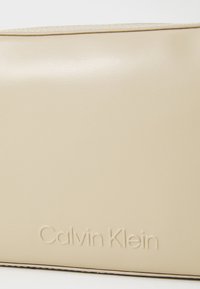 Calvin Klein šviesiai rudos odos pakuotė su lygiu paviršiumi, iškeltu logotipu ir kontrastinga siūle viršutiniame krašte. Stačiakampio formos.