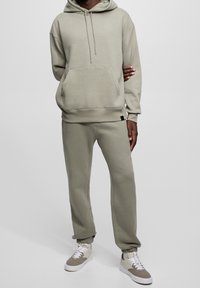 Sudadera con capucha gris claro y pantalones jogger a juego, hechos de tela suave, con bolsillo delantero y puños elásticos. Zapatillas deportivas con detalles en beige.