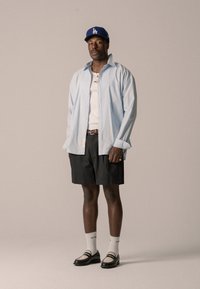 Hellblaue Button-Up-Hemd über einem weißen Tanktop, schwarze Shorts, weiße Socken und schwarz-braune Schuhe, gekrönt von einer blauen LA-Cap.