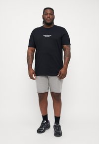 Sort bomulds T-shirt med hvid tekst, grå denim shorts, sorte sneakers og sorte sokker, stående mod en ensfarvet hvid baggrund.