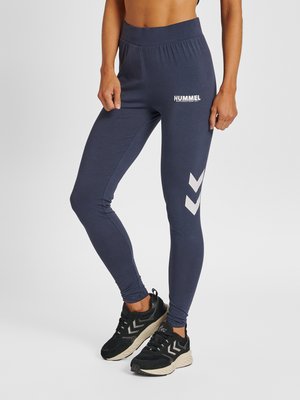 Vrouw draagt marineblauwe Hummel leggings met witte chevron-logo's en zwarte sportschoenen, staande tegen een effen achtergrond.