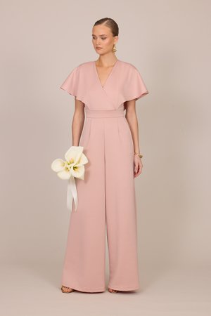 Frau trägt einen lachsrosa Jumpsuit mit weitem Bein und flatternden Ärmeln, hält weiße Calla-Lilien, die mit einem Band gebunden sind, und trägt goldene Accessoires.
