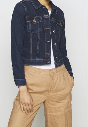 Person trägt eine dunkelblaue Jeansjacke mit goldenen Knöpfen über einem weißen Hemd und weite beige Hose mit einer Hand in der Tasche.