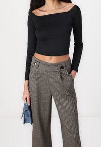 Schwarzes, schulterfreies Langarmoberteil kombiniert mit hochtaillierten Hosen in braun-schwarzem Hahnentritt mit zwei Knöpfen. Hält eine blaue Clutch.