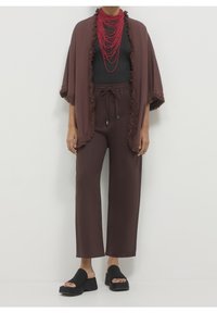 Personne portant un pantalon large marron foncé avec cordon, un châle à franges assorti, des sandales à plateforme noires à bout ouvert, un haut noir et un collier superposé de perles rouges.