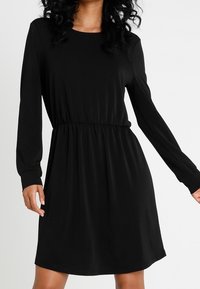 Svart långärmad klänning med rund halsringning, elastisk midja och slätt tyg, som har en lös passform och slutar ovanför knäna.