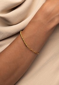 Pulsera de oro con una combinación de pequeñas cuentas y un diseño de cadena entrelazada, llevada en una muñeca, contra un fondo suave y neutral.