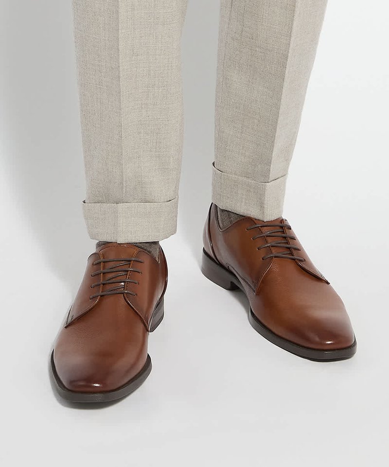 Chaussures habillées en cuir marron portées avec un pantalon beige clair retroussé et des chaussettes grises sur fond blanc.