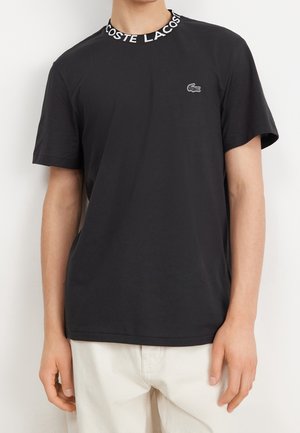 Černé bavlněné tričko s krátkými rukávy, s kulatým výstřihem, na kterém je bílé logo "LACOSTE" a malé logo Lacoste na hrudi.